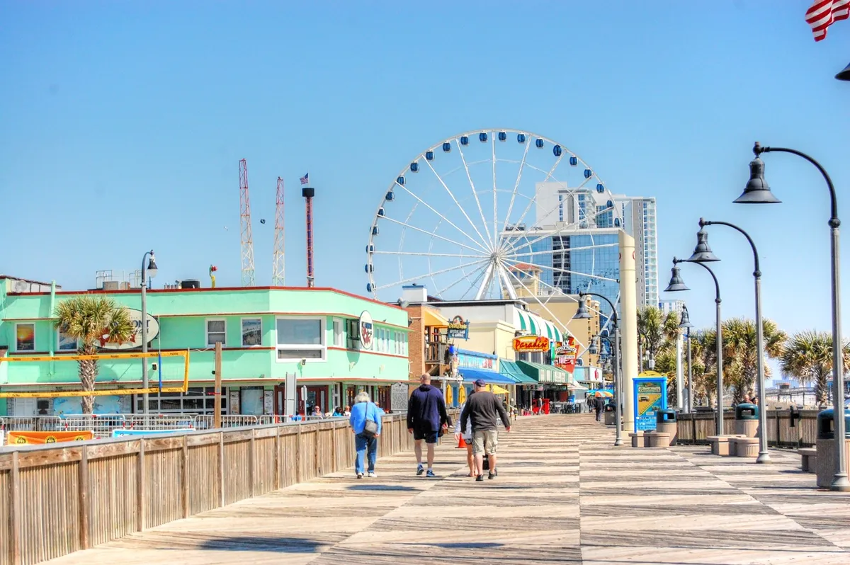 A lively exterior scene of Myrtle Beach's famous boardwalk, featuring the iconic Ferris wheel, colorful buildings, and people enjoying outdoor activities. The view captures both the city’s attractions and the relaxed energy of the area.