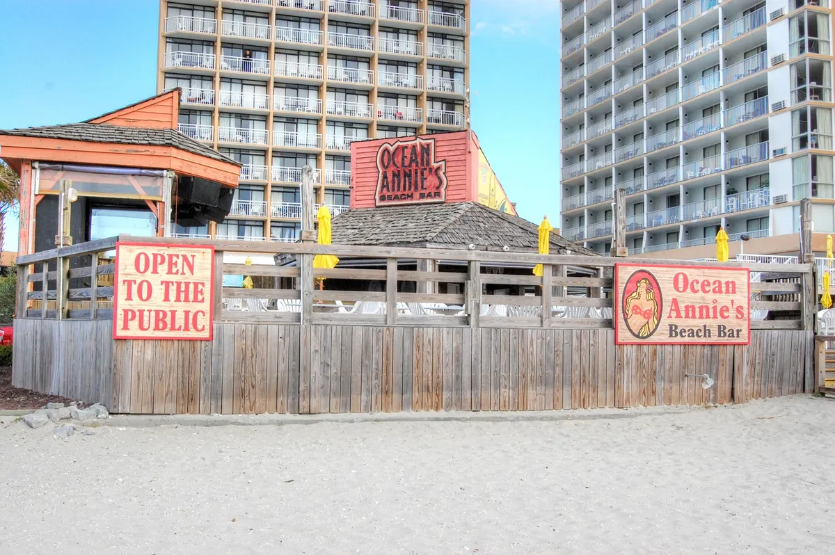 An inviting exterior shot of Ocean Annie's Beach Bar, with open signage, patio seating, and festive umbrellas. The scene buzzes with a relaxed, beachside energy, perfect for group hangouts, live music, and post-beach gatherings.