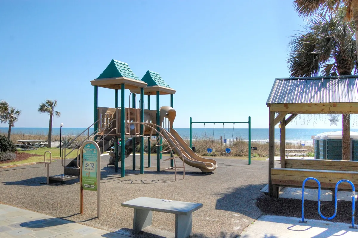 A lively playground set against a backdrop of palm trees and the shimmering ocean. Children play on swings and a playground structure, while families gather for picnics or strolls along the sandy beach. The setting radiates joy and vibrant family activity.