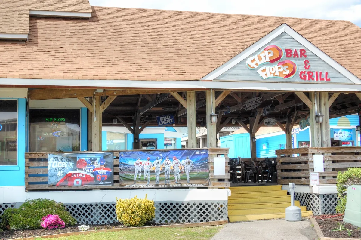 An exterior view of a lively local bar with sports banners, an inviting bar sign, and outdoor seating. The setup hints at an energetic environment ideal for post-game gatherings, dining with friends, or watching sports events in the open air.