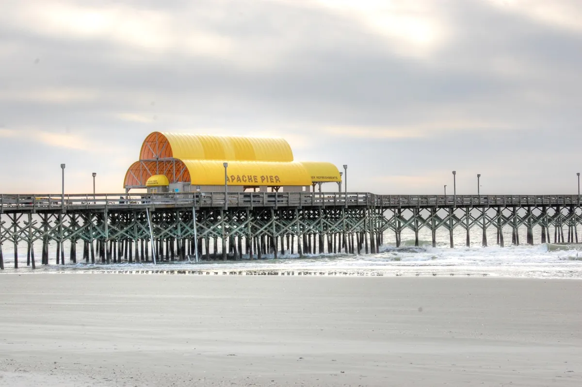 This image features the iconic Apache Pier stretching out into the ocean, with its distinctive yellow roof and sturdy wooden structure. The sandy shoreline in the foreground invites families to imagine walks at dawn, sandcastle-building, and gentle waves perfect for kids and parents alike.