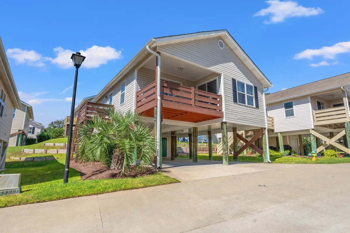 A cheerful exterior view of the house featuring a spacious balcony, palm trees, and a driveway leading up to the home. The balcony is prominent, suggesting opportunities for enjoying the coastal atmosphere from an elevated vantage point.