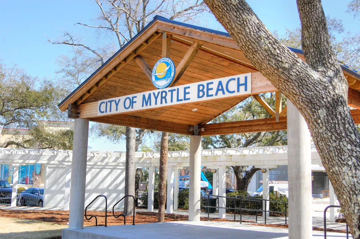 An exterior view featuring the 'City of Myrtle Beach' sign, a charming wooden pavilion structure, and an inviting landscape of surrounding trees. The space is clearly set up for community gatherings and public events, providing a glimpse of the welcoming neighborhood atmosphere.