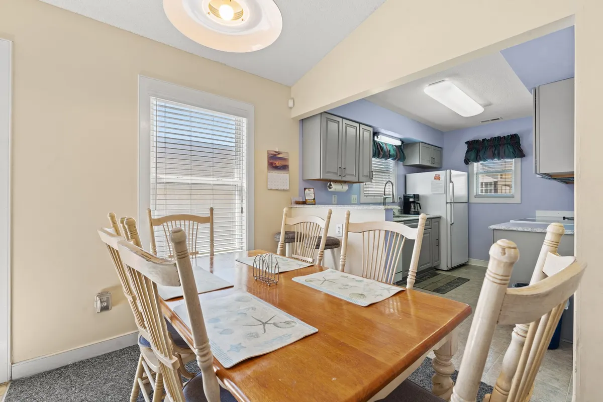 A welcoming dining area connected to a modern kitchen, featuring a dining table with ample seating, a kitchen area in the background, and sunlight streaming through a window. The setup encourages family meals, celebrations, and communal activities.