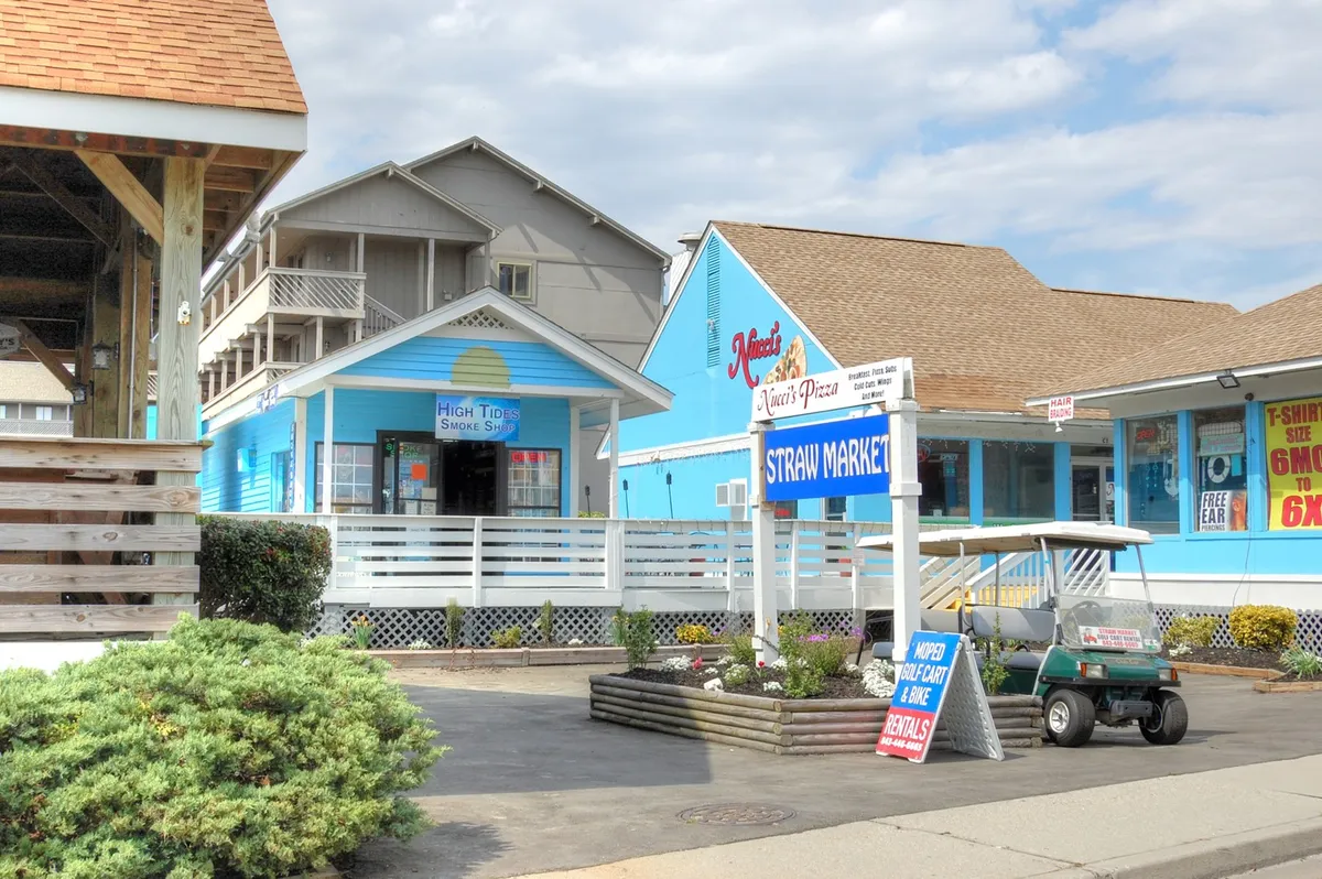 A vibrant exterior shot featuring colorful buildings, a sign for the Straw Market, a golf cart parked nearby, and an inviting outdoor seating area. The atmosphere is lively and relaxed, ideal for exploring local shops and eateries or renting equipment for more adventures.