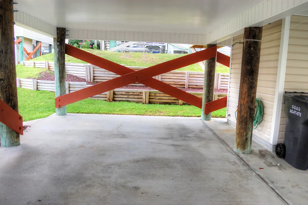 A practical and spacious covered parking area beneath the raised house, with cross-braced wooden supports and concrete flooring. Greenery is visible through the open sides, blending function with a connection to the outdoors.