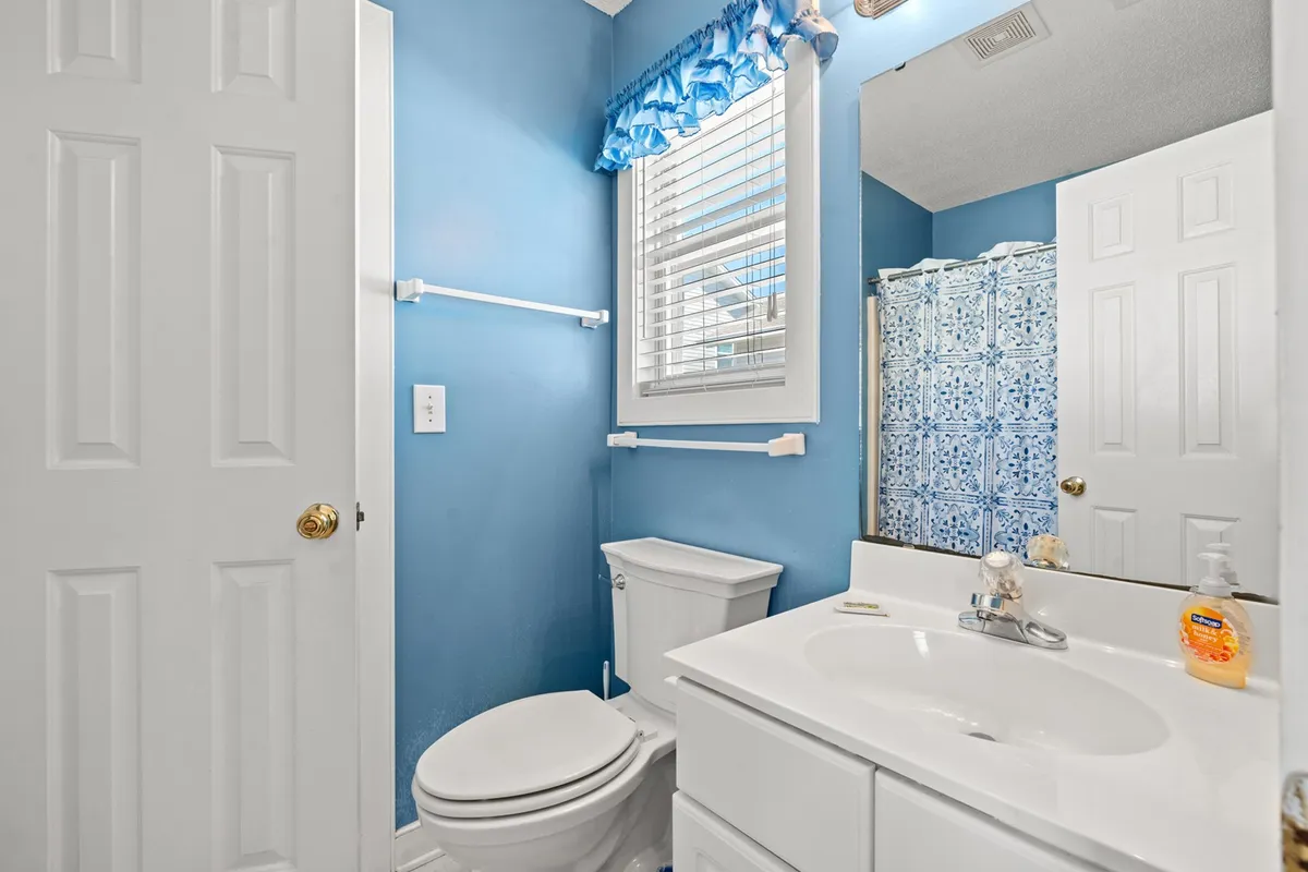 A fresh and clean bathroom with blue walls, white fixtures, a decorative shower curtain, and natural light streaming through blinds. The space feels inviting and well-maintained, suitable for both quick routines and relaxation.