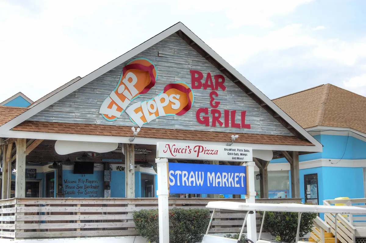 A lively scene at the Flip Flops Bar & Grill, with visible signage for both the bar and Nucci's Pizza alongside the Straw Market. The setting showcases outdoor dining and local eateries, emphasizing the diversity of dining and shopping options available nearby.